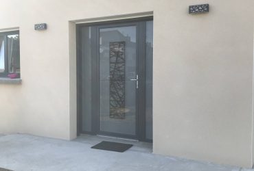 Porte d'entrée vitrée modèle Logan disposant d'un vitrage dépoli, de deux tierces fixes vitrées assorties, le tout dans un coloris Gris anthracite RAL 7016. Elle fut installée avec des fenêtres SO finition extérieure alu 7016 et finition intérieure PVC blanc